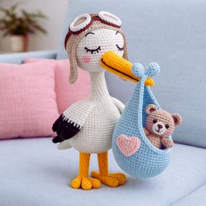 Puede incluir: Una cigüeña de ganchillo hecha a mano con gorro de piloto, sosteniendo una bolsa azul con un osito de peluche. La cigüeña tiene pico y patas amarillas, con alas negras y blancas. Un corazón rosa está en la bolsa.