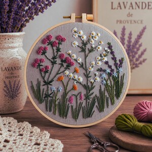 May include: A circular wooden embroidery hoop containing grey fabric with hand-stitched wildflowers in shades of pink, white, orange, blue, and purple. The design features various stems and green leaves, secured by a metal screw at the top of the frame.