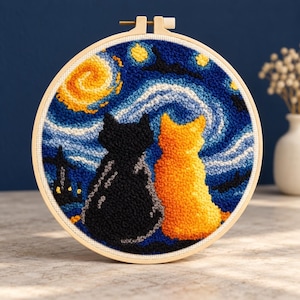 Pode incluir: Um bastidor de bordado circular de madeira contendo uma peça de arte em punch needle. O design apresenta um gato preto e um gato laranja sentados lado a lado, de frente para um céu noturno azul com uma lua amarela e estrelas amarelas.