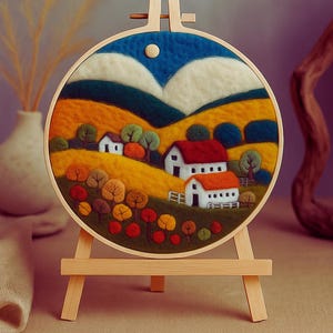 May include: A circular felt landscape artwork in a wooden embroidery hoop. The scene shows white houses with red roofs, and colourful trees. The background features rolling hills and a blue sky with a white moon. The hoop is displayed on a small wooden easel.