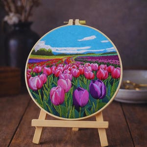Può includere: Ricamo a mano di un campo di tulipani in fiore, presentato in un telaio di legno rotondo su un piccolo cavalletto di legno. Il cielo è blu con nuvole bianche, e i tulipani sono rosa e viola. Un'opera d'arte decorativa.