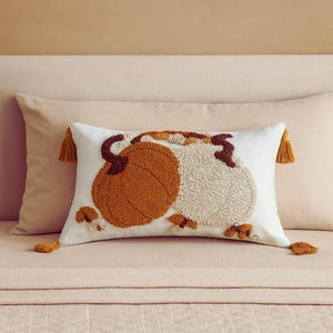 Könnte beinhalten: Weißes rechteckiges Kissen mit zwei gestickten Kürbissen in Orange und Creme. Das Kissen hat braune Stiele und Akzente, mit goldenen Quasten an jedem Ende. Ein herbstliches Akzent für ein Bett oder Sofa.