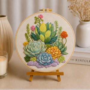Könnte beinhalten: Ein runder Stickrahmen mit einem farbenfrohen Sukkulenten-Design. Das Kunstwerk zeigt verschiedene Sukkulenten in Grün-, Gelb-, Orange- und Blautönen mit rosa Akzenten. Die Stickerei wird auf einer kleinen Holzstaffelei präsentiert.