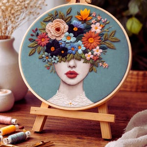 Può includere: Opera d'arte ricamata di un volto femminile con un copricapo floreale, in un telaio di legno su un piccolo cavalletto di legno. La donna ha labbra rosse e un colletto di pizzo bianco. Bobine di filo colorate e forbici in primo piano, con misure in cm.