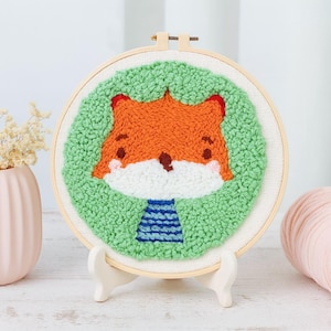May include: A handmade embroidery hoop art piece featuring a cartoon fox. The fox has orange fur, red ears, and a white muzzle, surrounded by green textured stitching. The fox wears a blue and white striped shirt. The artwork is displayed on a wooden stand.