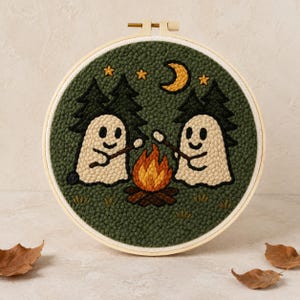 Peut inclure: Un tambour à broder circulaire contenant une œuvre en punch needle représentant deux fantômes blancs faisant griller des guimauves sur un feu de camp. Le décor inclut des sapins vert foncé, un croissant de lune jaune et des étoiles sur fond vert.
