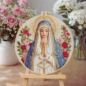 Può includere: Opera d'arte ricamata di una donna in una veste blu e bianca con le mani giunte in preghiera, circondata da un'aureola dorata e rose. L'opera è esposta su un piccolo cavalletto di legno. Rose rosa e fiori bianchi sullo sfondo.