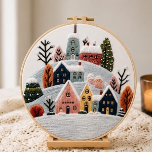 May include: A circular embroidery hoop containing a fabric panel with a hand-stitched winter village scene. The design features colorful houses in pink, yellow, blue, and red, surrounded by snow-covered hills, bare trees, and evergreen pines.