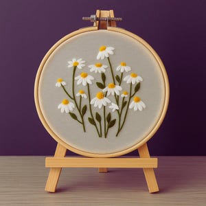 Puede incluir: Un bastidor de bordado redondo con un diseño floral de margaritas. Las margaritas tienen pétalos blancos y centros amarillos, con tallos y hojas verdes. El bordado está sobre una tela gris claro, exhibido en un pequeño caballete de madera.