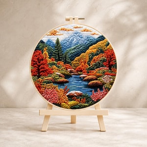 May include: A circular embroidery hoop containing a detailed landscape scene of a blue mountain range, a winding river, and dense autumn foliage in shades of red, orange, and yellow, all mounted on a small, light-colored wooden display easel.