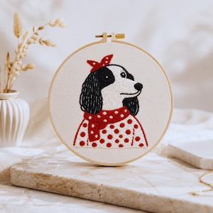 Può includere: Opera d'arte ricamata in un cerchio di legno, raffigurante un cane bianco e nero con una sciarpa e un fiocco a pois rossi. Il cane è ricamato su tessuto color crema. Il pezzo ha una qualità artigianale, con un design affascinante.