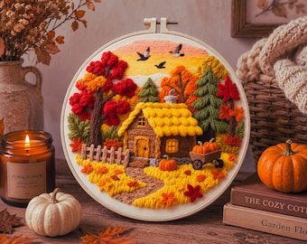 May include: A circular embroidery hoop featuring a textured, three-dimensional landscape of a cozy cabin in autumn. The scene includes vibrant red and orange trees, a yellow-roofed house, a small wooden cart with pumpkins, and birds in flight.