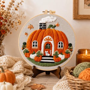 Pode incluir: Um bastidor de bordado circular com um design costurado de uma grande casa de abóbora laranja com porta branca, janelas e uma chaminé com fumaça. Um pequeno gato branco senta-se na porta, rodeado por abóboras menores e folhagem verde.