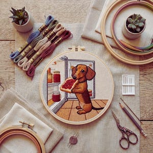 May include: An embroidered hoop art piece depicting a dachshund with toast in front of a fridge. The image includes embroidery threads, hoops, a small succulent plant, and other craft supplies, all arranged on a neutral fabric.
