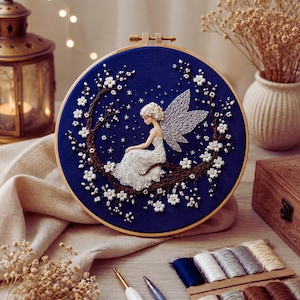 Peut inclure: Un tambour à broder circulaire présentant un tissu bleu profond avec un motif cousu à la main représentant une fée aux ailes blanches assise sur une branche brune. La branche est ornée de petites fleurs blanches et de petites étoiles.