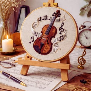 Puede incluir: Un violín bordado con notas musicales sobre tela color crema, en un aro de madera. La obra de arte se exhibe en un pequeño caballete de madera. También se ve una figura de clave de sol y partituras.