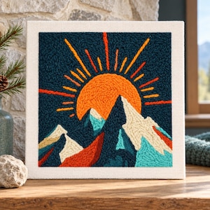 Könnte beinhalten: Ein quadratisches Punch-Needle-Wandbild mit texturiertem Landschaftsmotiv. Das Bild zeigt eine leuchtend orangefarbene Sonne, die über dunkelblauen Bergen mit weißen und hellblauen schneebedeckten Gipfeln vor einem dunkelblauen Himmel aufgeht.