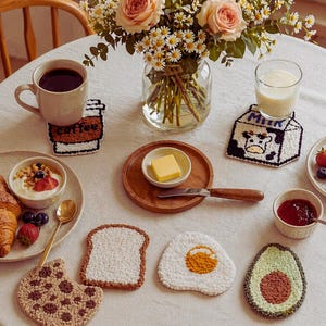 Puede incluir: Una mesa de desayuno con una taza de café, leche y varios alimentos. Incluye un cruasán, yogur con bayas y posavasos con forma de taza de café, cartón de leche, tostada, huevo, aguacate y galleta. Un jarrón de flores está en el fondo.