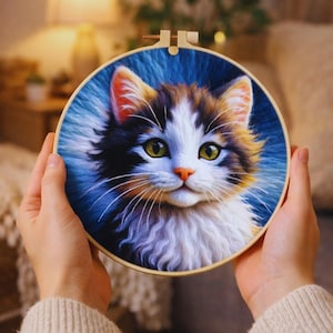 May include: A colorful cat embroidery in a wooden hoop. The cat has white, brown, and black fur with green eyes and an orange nose. The background is a gradient of blue and white. The embroidery is held by a person.