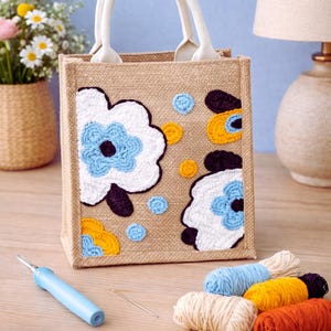 Op de afbeelding: Een beige jute draagtas met witte handvatten, versierd met geborduurde witte, blauwe, gele en zwarte bloemmotieven. Knutselbenodigdheden, waaronder garen en een naaldgereedschap, zijn in de buurt gerangschikt. De tas is een handgemaakt item.