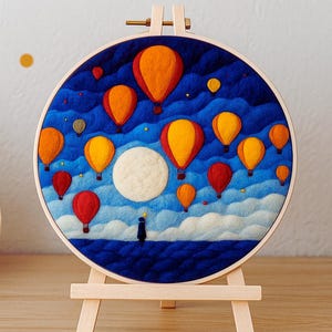 Könnte beinhalten: Ein rundes Filz-Kunstwerk, das eine Nachtlandschaft mit roten, orangefarbenen und gelben Heißluftballons zeigt. Ein weißer Mond und eine Figur sind ebenfalls abgebildet. Das Kunstwerk wird auf einer kleinen Holzstaffelei präsentiert.