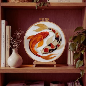 Op de afbeelding: Geborduurde koi-viskunst in een houten hoepel. Het kunstwerk toont twee koi-vissen, één oranje en één zwart-wit, op een witte achtergrond. Het stuk wordt tentoongesteld op een kleine houten ezel op een houten plank.