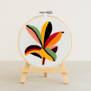 Puede incluir: Diseño de hoja bordada en un aro de madera. La hoja presenta tonos verdes, amarillos, rojos, negros y rosas. El bordado se exhibe en un pequeño caballete de madera, creando una pieza decorativa.