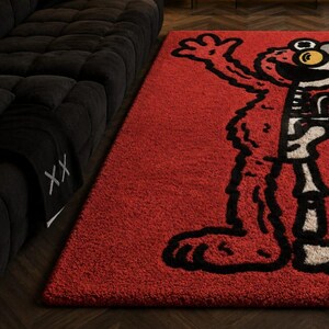 Puede incluir: Una alfombra rectangular roja con un dise&ntilde;o de personaje de dibujos animados en blanco y negro. La alfombra est&aacute; colocada sobre un suelo de madera oscura junto a un sof&aacute; negro. El dise&ntilde;o de la alfombra es un personaje estilizado con una cara grande y expresiva.