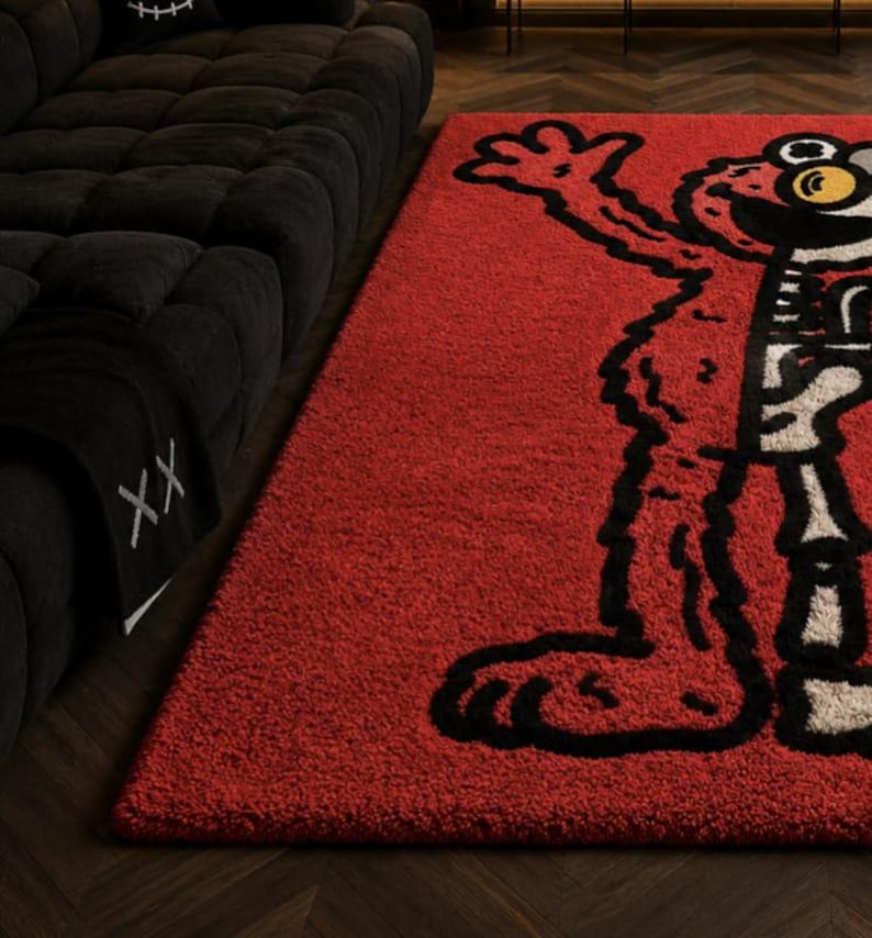 Puede incluir: Una alfombra rectangular roja con un dise&ntilde;o de personaje de dibujos animados en blanco y negro. La alfombra est&aacute; colocada sobre un suelo de madera oscura junto a un sof&aacute; negro. El dise&ntilde;o de la alfombra es un personaje estilizado con una cara grande y expresiva.