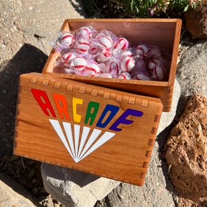 May include: A wooden box with the word "ARCADE" in rainbow colors and a white fan design. The box is open, revealing individually wrapped red and white striped mint candies. The box is made of light brown wood.