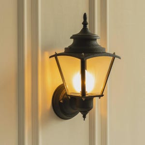 Peut inclure: Applique murale d'extérieur noire avec une lumière jaune chaude. La lanterne a un design classique avec un toit pointu et des panneaux de verre transparents. Le luminaire est monté sur une base ronde.