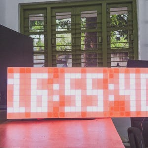 May include: A digital clock displaying the time 16:55:40. The clock face is a grid of orange and white squares, with the numbers illuminated. The clock is on a red surface, with a dark monitor and other objects in the background.