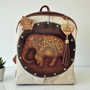 Puede incluir: Mochila de cuero artesanal con un diseño de elefante en relieve. La mochila presenta un círculo de cuero marrón con un elefante en relieve, rodeado de un patrón de piel de vaca. Un dije de corazón de madera con el texto "I LOVE ARTESANAL" cuelga de la cremallera.
