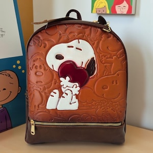 May include: A brown Snoopy backpack with a gold zipper and a brown base. The front features an embossed Snoopy holding a red heart, with other embossed Peanuts characters. The backpack has a top handle and adjustable straps.