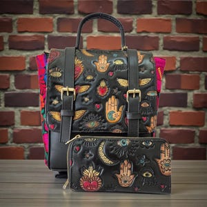 Puede incluir: Conjunto de mochila y cartera de cuero negro. La mochila tiene hebillas doradas, un asa superior y correas ajustables. Ambos artículos están grabados con diseños coloridos de corazón sagrado, ojo, mano y alas. La mochila tiene un forro textil colorido.