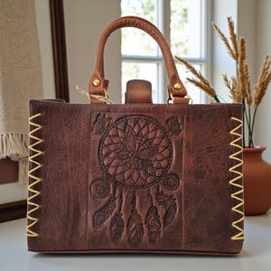 Puede incluir: Bolso tote de cuero marrón con un diseño de atrapasueños en relieve en la parte delantera. El bolso tiene un asa superior, un cierre de solapa y costuras doradas en los bordes. El bolso rectangular parece estar hecho de cuero envejecido.