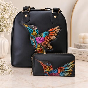 Puede incluir: Una mochila negra y una cartera rectangular a juego, ambas con un diseño de colibrí multicolor vibrante con patrones intrincados en tonos azul, naranja, amarillo y rosa, acentuadas con herrajes y cremalleras en tono dorado.