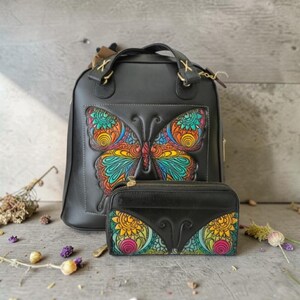 May include: Black leather backpack and wallet set. The backpack features a large, colorful butterfly design on the front. The wallet has a matching butterfly wing design. The butterfly designs are in shades of blue, yellow, orange, and red.