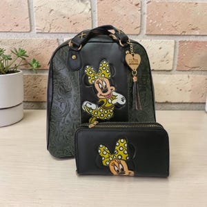 Op de afbeelding: Zwarte rugzak- en portemonneeset met Minnie Mouse. De rugzak heeft een bloemreli&euml;f en goudkleurige hardware. De portemonnee en rugzak hebben beide een Minnie Mouse applicatie met een gele stippen strik.