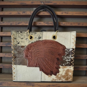 Puede incluir: Bolso tote patchwork con un diseño de jefe nativo americano de cuero marrón. El bolso presenta una mezcla de patrones de piel de vaca en blanco, marrón y negro. Tiene asas negras y aros metálicos dorados.