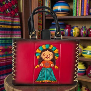 Puede incluir: Un bolso rojo con asa negra y diseño de muñeca decorativa. La muñeca tiene un tocado colorido, top naranja y falda azul. El bolso tiene un borde cosido y está sobre una superficie de madera.
