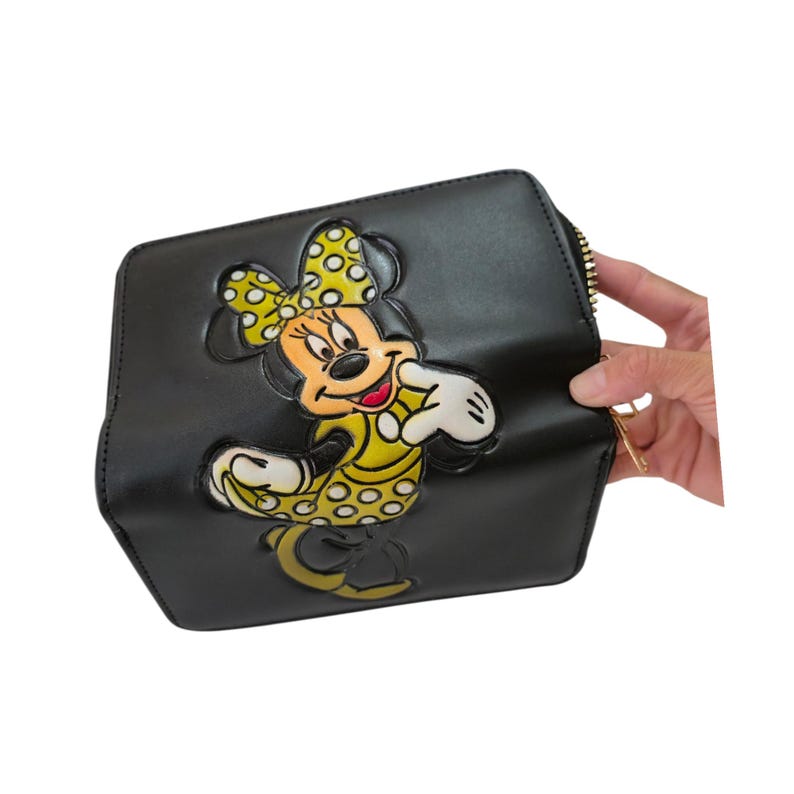 Op de afbeelding: Zwarte clutch met een gele Minnie Mouse-ontwerp met stippen. Het stripfiguur draagt een gele jurk en strik. De clutch heeft een gouden rits en een rechthoekige vorm.