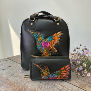 Puede incluir: Conjunto de mochila y cartera negras. Ambos artículos presentan un diseño de colibrí colorido con detalles en naranja, azul, amarillo y rosa. La mochila tiene un asa superior y herrajes dorados. La cartera es de estilo cremallera.