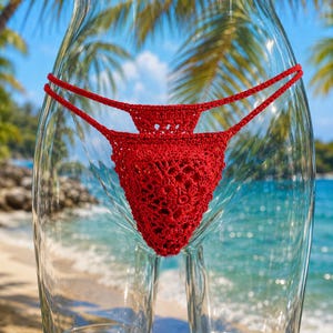 Könnte beinhalten: Ein leuchtend rotes Häkel-Bikiniunterteil mit einem komplizierten, blumenartigen Spitzenmuster. Das Kleidungsstück ist mit dünnen, verstellbaren seitlichen Bindebändern versehen, die um die Hüften verlaufen und eine feine, detaillierte Textur erzeugen.