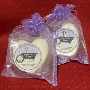 May include: Two heart-shaped goat milk soaps in clear organza bags tied with lavender ribbon. Each soap has a round label with the text "Goat Milk Soap" and "Lavender". The ingredients are listed below the text. The background is a red surface.