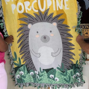 May include: A yellow fleece blanket with a cartoon hedgehog design. The blanket features the text "RISE AND SHINE PORCUPINE" in white letters. The hedgehog is gray with a white belly and pink cheeks. The blanket has a green and gray fringe.