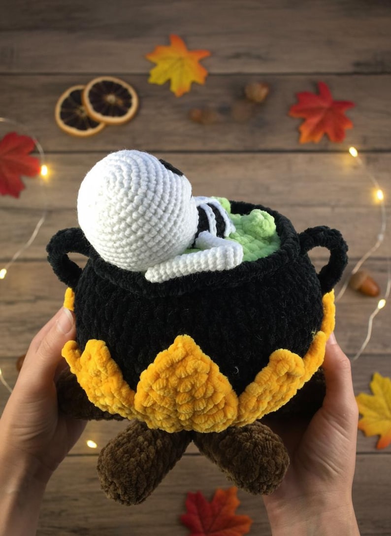 Crochet Halloween amigurumi pattern featuring a black plush cauldron with yellow flames, a bubbling green potion, and a cute white skeleton sitting inside. Perfect for spooky home décor, Halloween party centerpiece, or a handmade gift. This detailed crochet pattern is ideal for amigurumi lovers and includes step-by-step instructions. Great project for Halloween crafters looking for unique and fun holiday decorations.