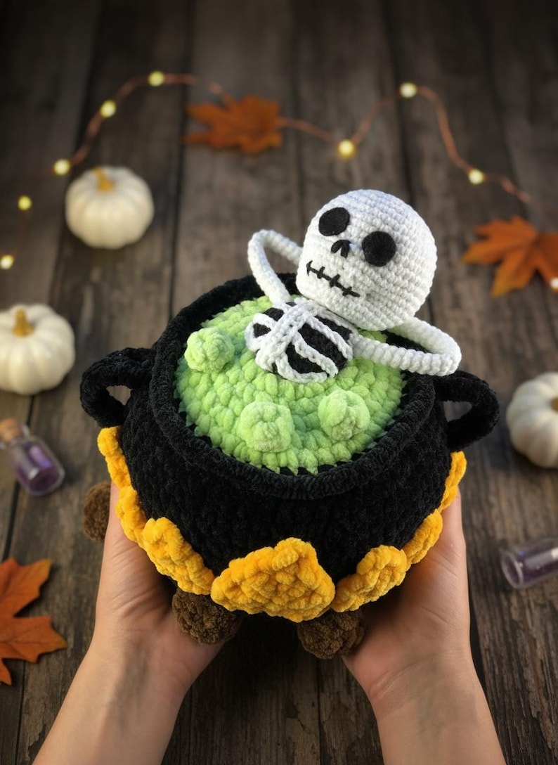 Crochet Halloween amigurumi pattern featuring a black plush cauldron with yellow flames, a bubbling green potion, and a cute white skeleton sitting inside. Perfect for spooky home décor, Halloween party centerpiece, or a handmade gift. This detailed crochet pattern is ideal for amigurumi lovers and includes step-by-step instructions. Great project for Halloween crafters looking for unique and fun holiday decorations.