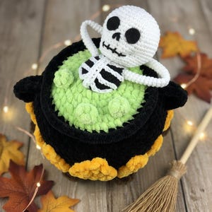 Crochet Halloween amigurumi pattern featuring a black plush cauldron with yellow flames, a bubbling green potion, and a cute white skeleton sitting inside. Perfect for spooky home décor, Halloween party centerpiece, or a handmade gift. This detailed crochet pattern is ideal for amigurumi lovers and includes step-by-step instructions. Great project for Halloween crafters looking for unique and fun holiday decorations.