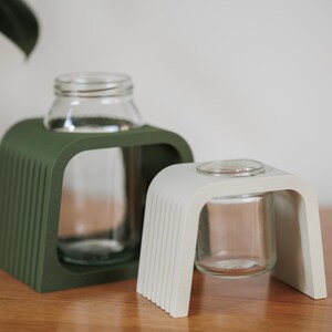 May include: Two decorative glass bottle holders. One is a dark green, the other is off-white. Each holder has a curved, arched design with a clear glass bottle inside. The holders are made of a solid material with vertical lines on the front.
